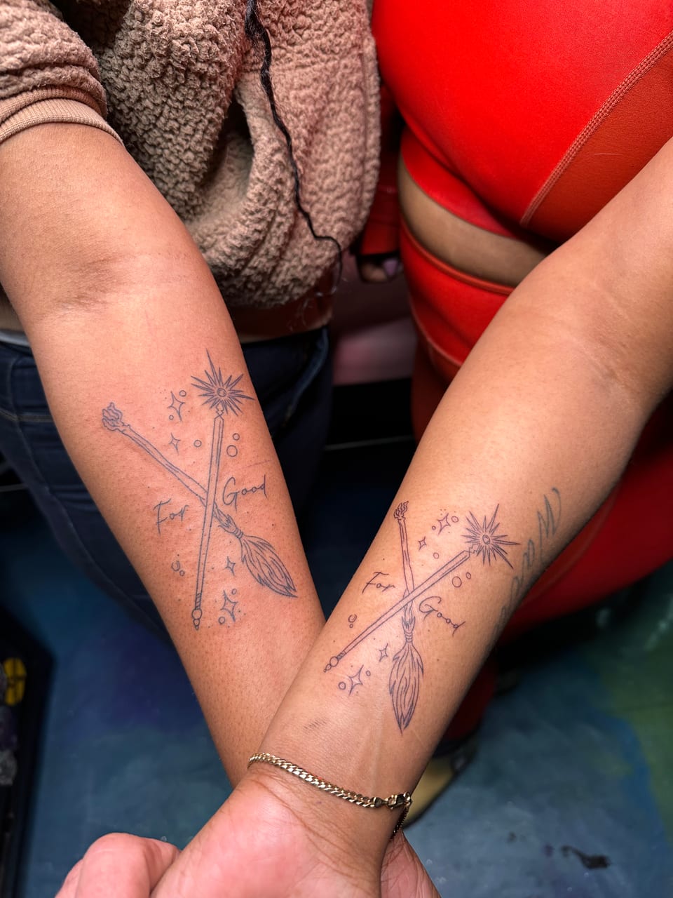 Matching fine-line wand and broom tattoos cross the words For Good on forearms.