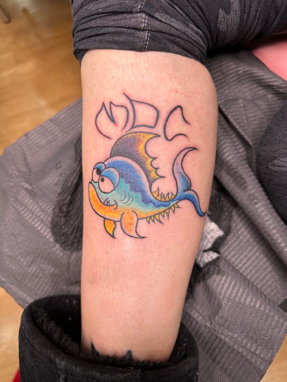 A blue and orange cartoon fish tattoo sits beneath outlined lettering on a forearm.