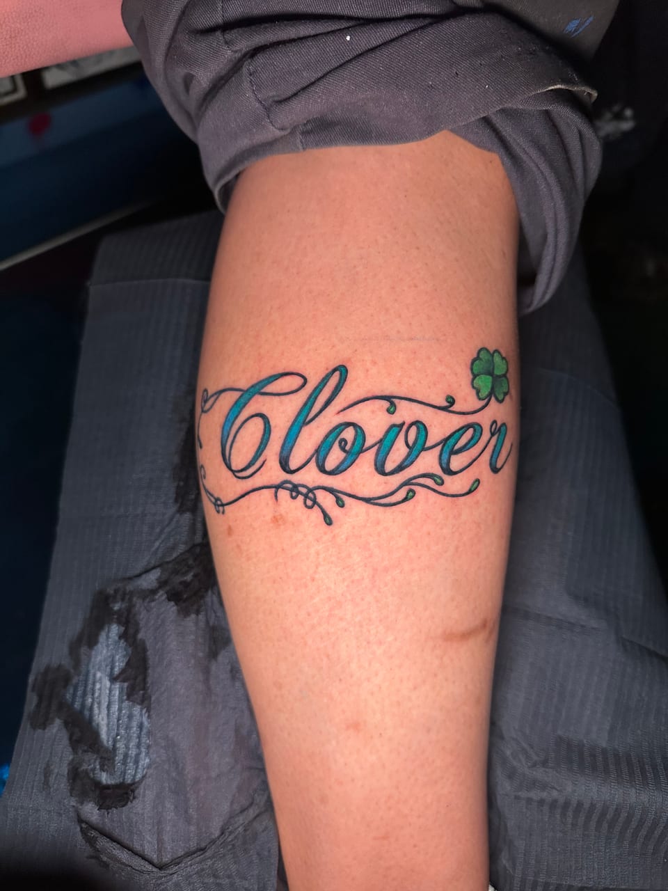 Teal cursive lettering and a green clover tattoo wrap across a forearm.