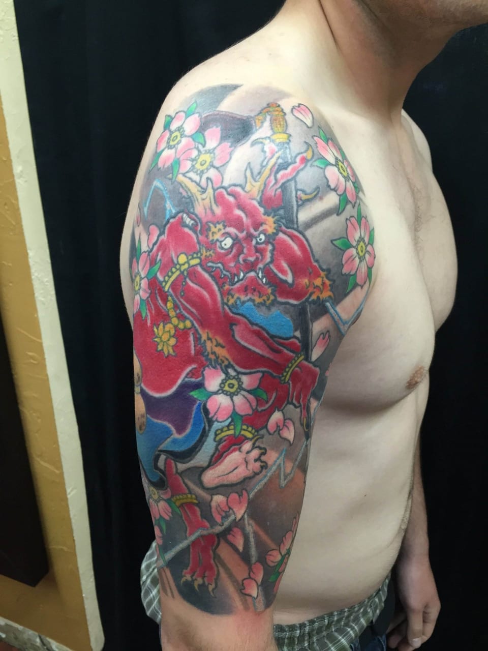 Colorful tattoo of a red demon with horns, surrounded by pink cherry blossoms on the upper arm.