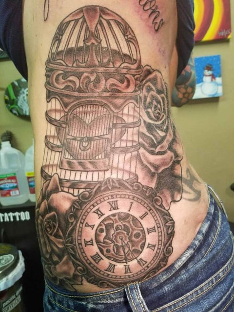 A black and grey birdcage, roses and a pocket watch run down the ribs to a hip.