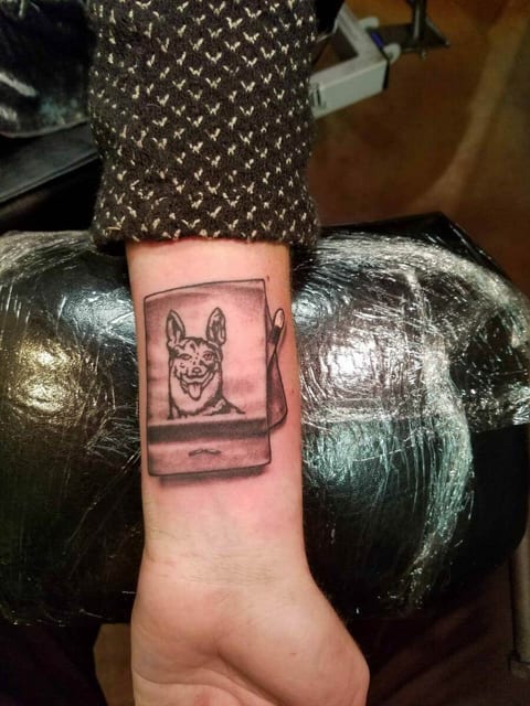 A black and grey illustrative dog inside an instant-print frame sits on an inner forearm.