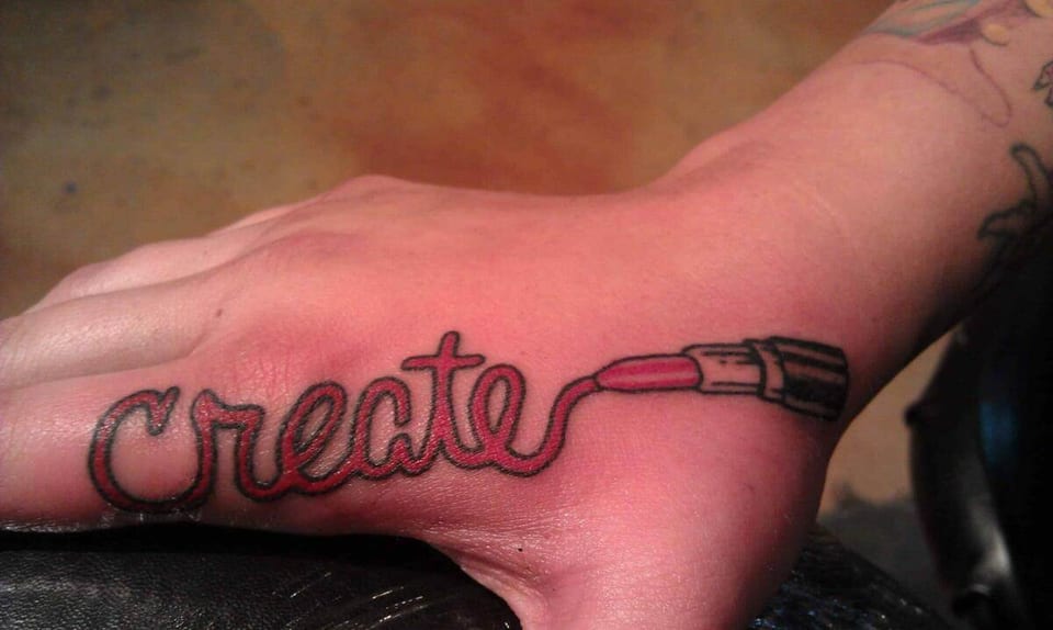 Red script lettering reading 'Create' trails into a lipstick tattoo on the back of a hand.