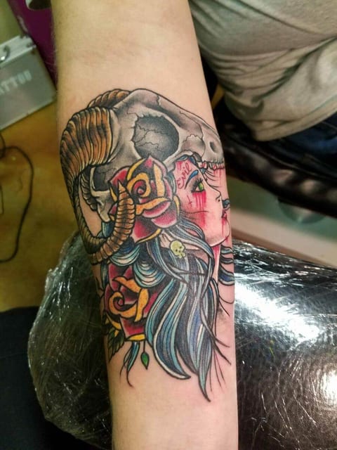 A traditional color woman with blue hair, a ram skull and roses covers a forearm.