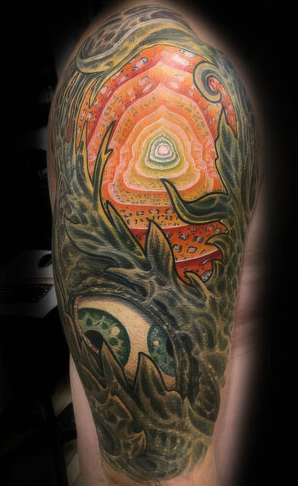 A color bio-organic tattoo with orange concentric shapes and green eyes covers an upper arm.
