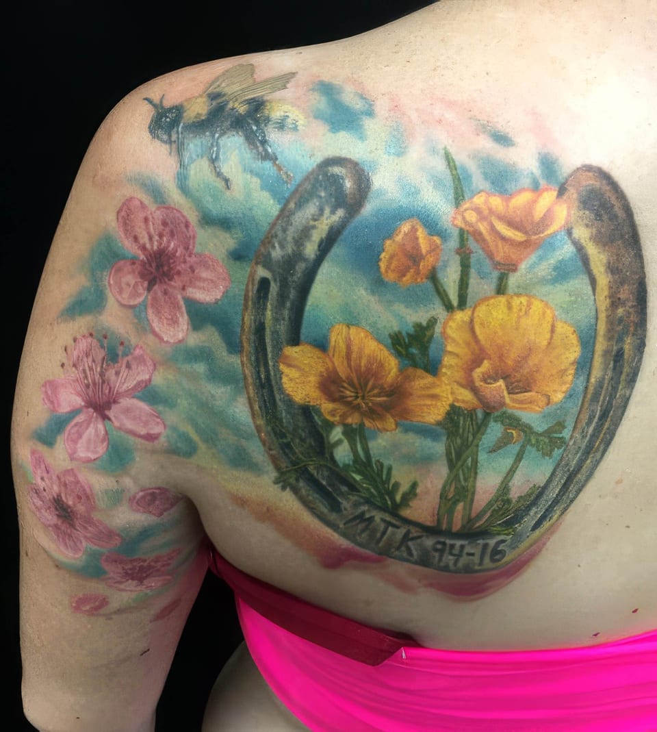 A colorful tattoo on a person's upper back featuring pink flowers, orange poppies, a horseshoe, and a bee.