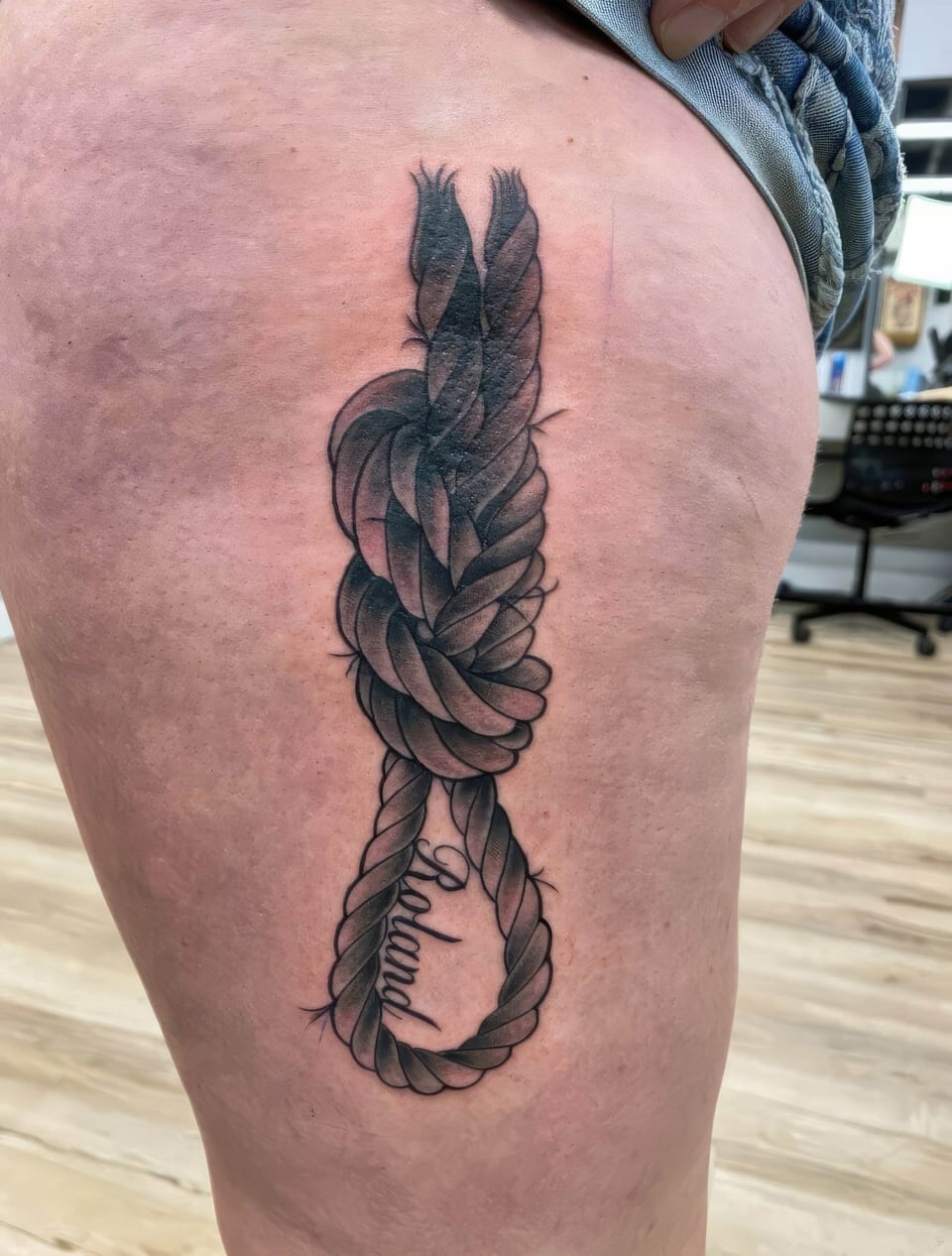 A black and grey illustrative rope knot and loop surround script on a thigh.