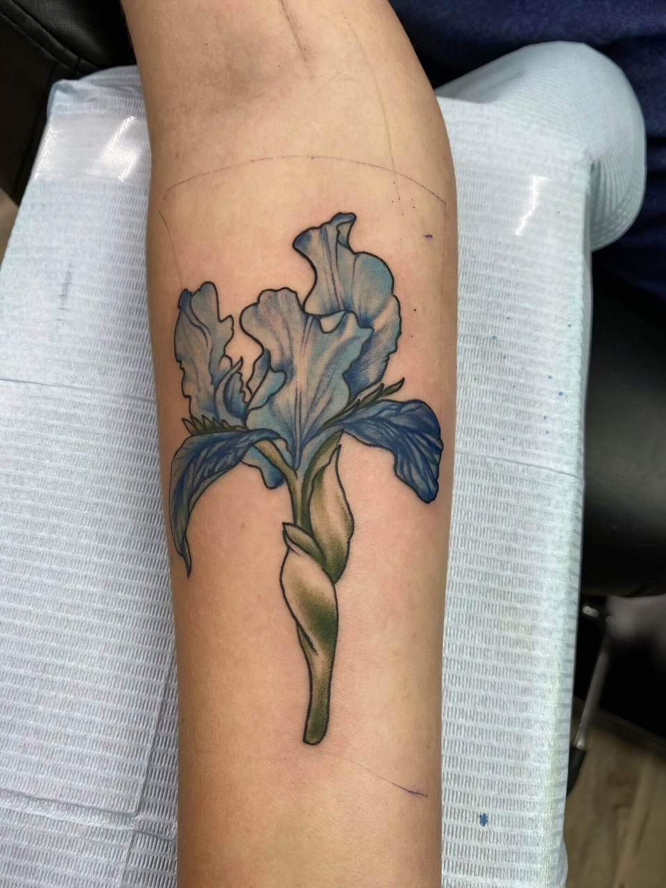 A realistic blue iris with a green stem covers an inner forearm.
