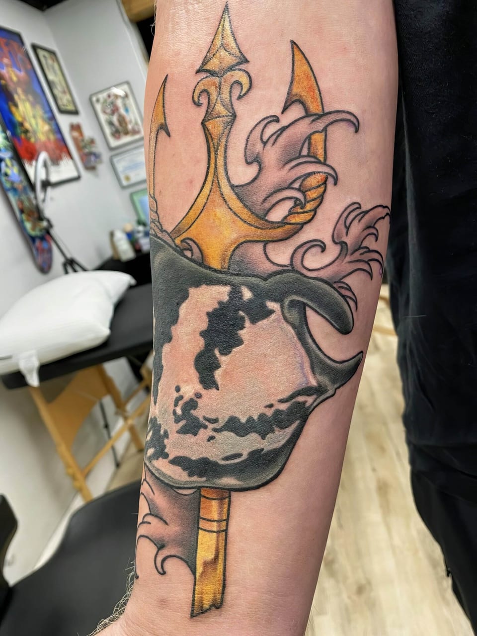 An angled view shows a color illustrative stingray, golden trident and waves tattooed on the forearm beside a pillow.