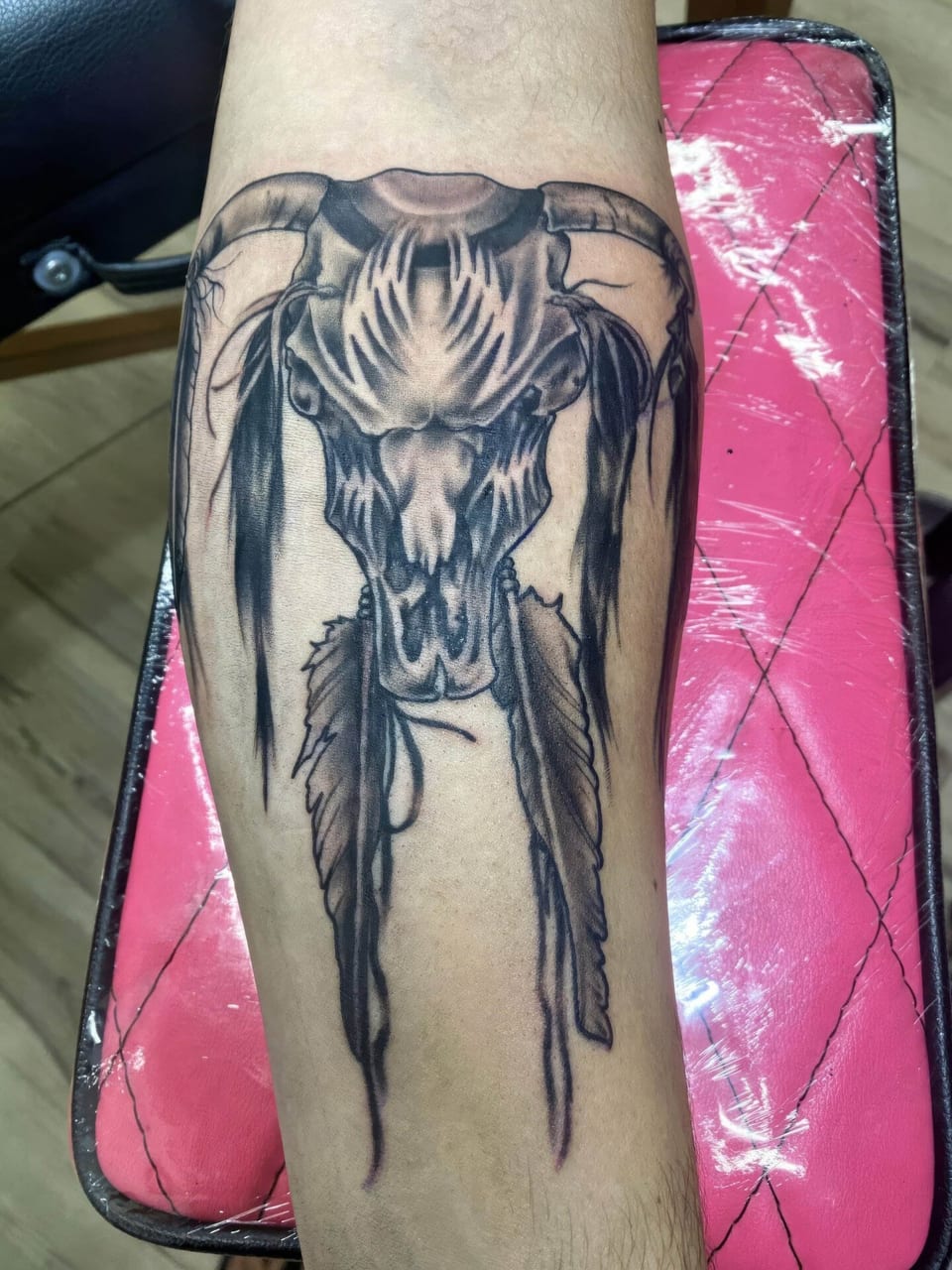 A black and grey illustrative horned skull with hanging feathers covers a forearm.