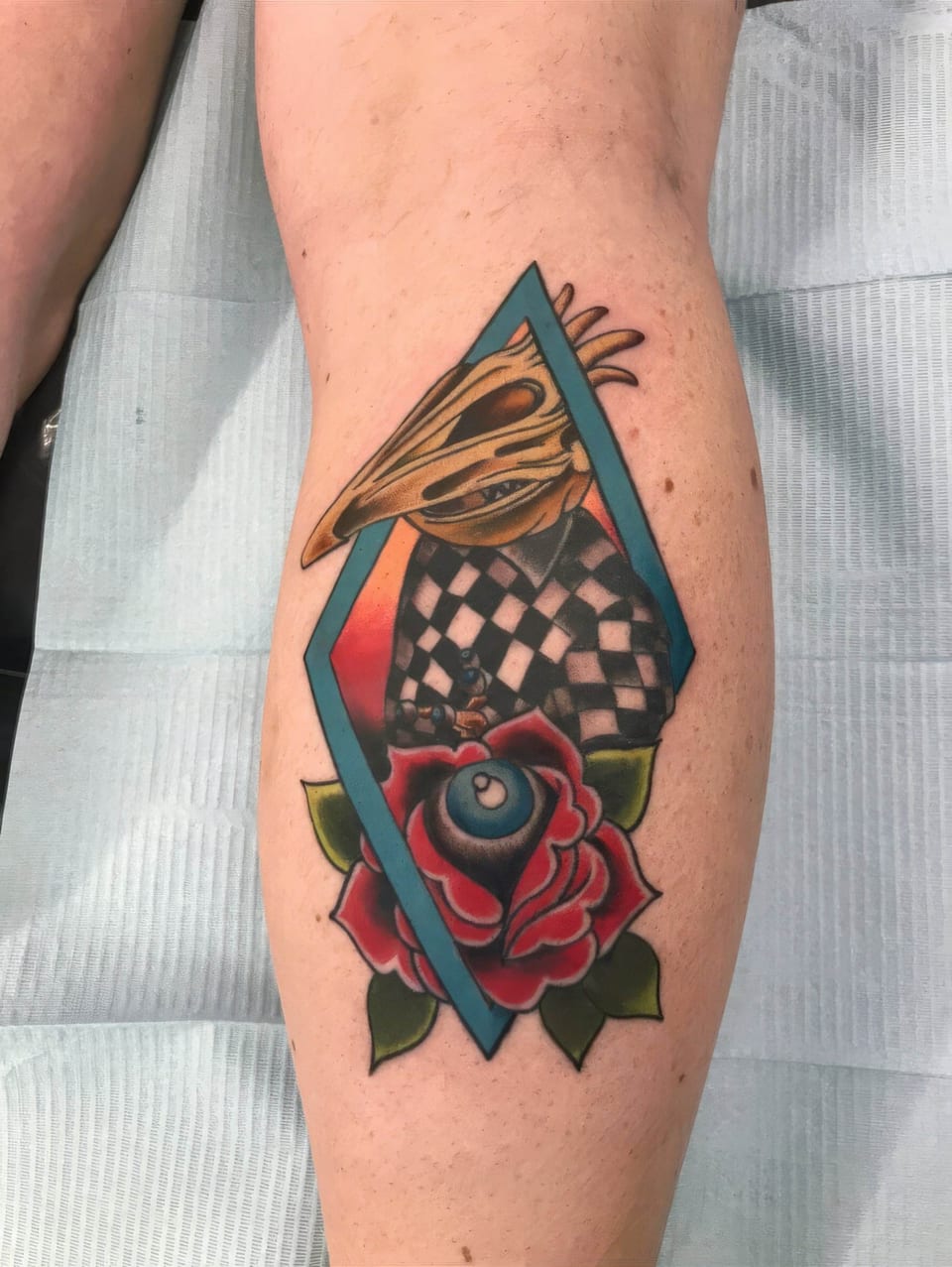 A colorful illustrative bird skull, checkerboard and rose sit within a diamond frame on the calf.
