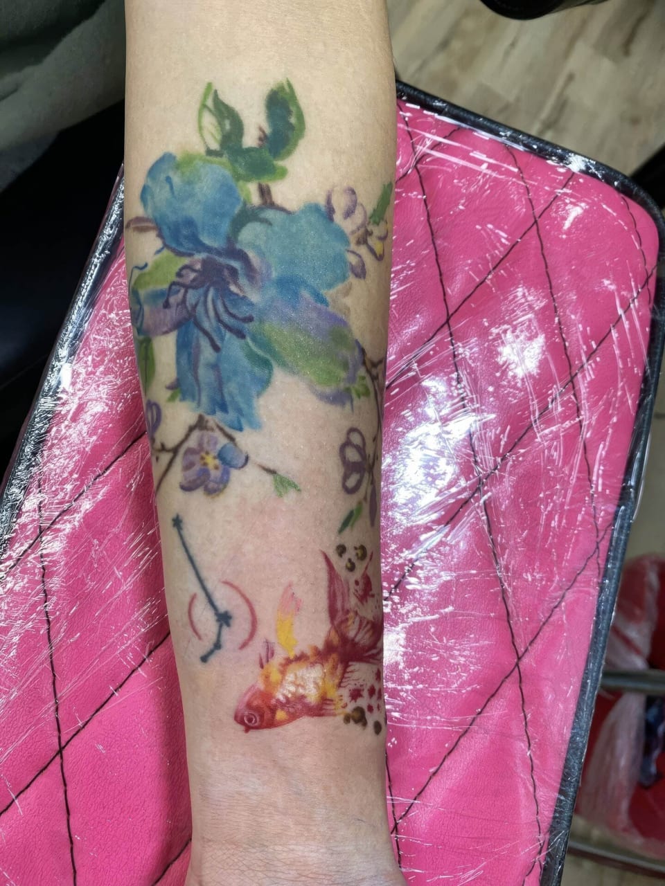 Watercolor blue flowers and a red and gold fish run along the forearm toward the wrist.
