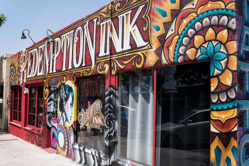 Redemption Ink's storefront displays painted lettering, a woman's portrait beside the door and a mandala mural at right.