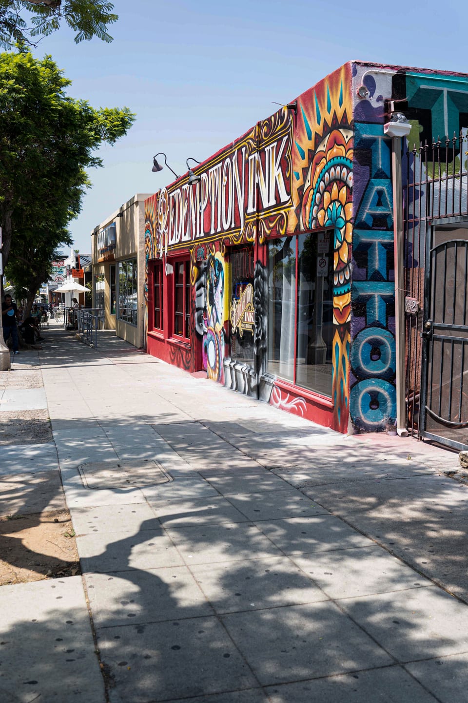 Sidewalk view down the block toward the Redemption Ink building, with its lettering, teal TATTOO letters and a black gate.