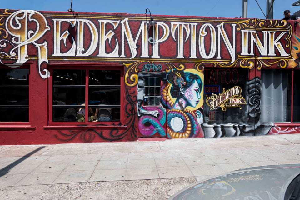 Closest front view of the Redemption Ink lettering, portrait mural, Open sign and window decal, with a car hood below.