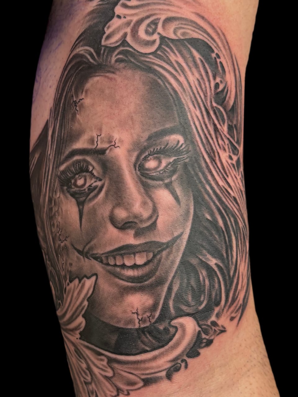 A black and grey realistic woman with clown makeup, cracked skin and a wide grin covers an outer upper arm.