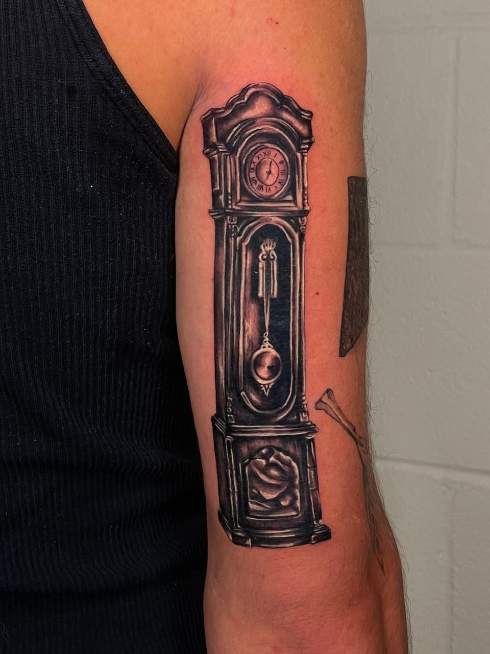 A black and grey grandfather clock tattoo with a pendulum runs down an arm.